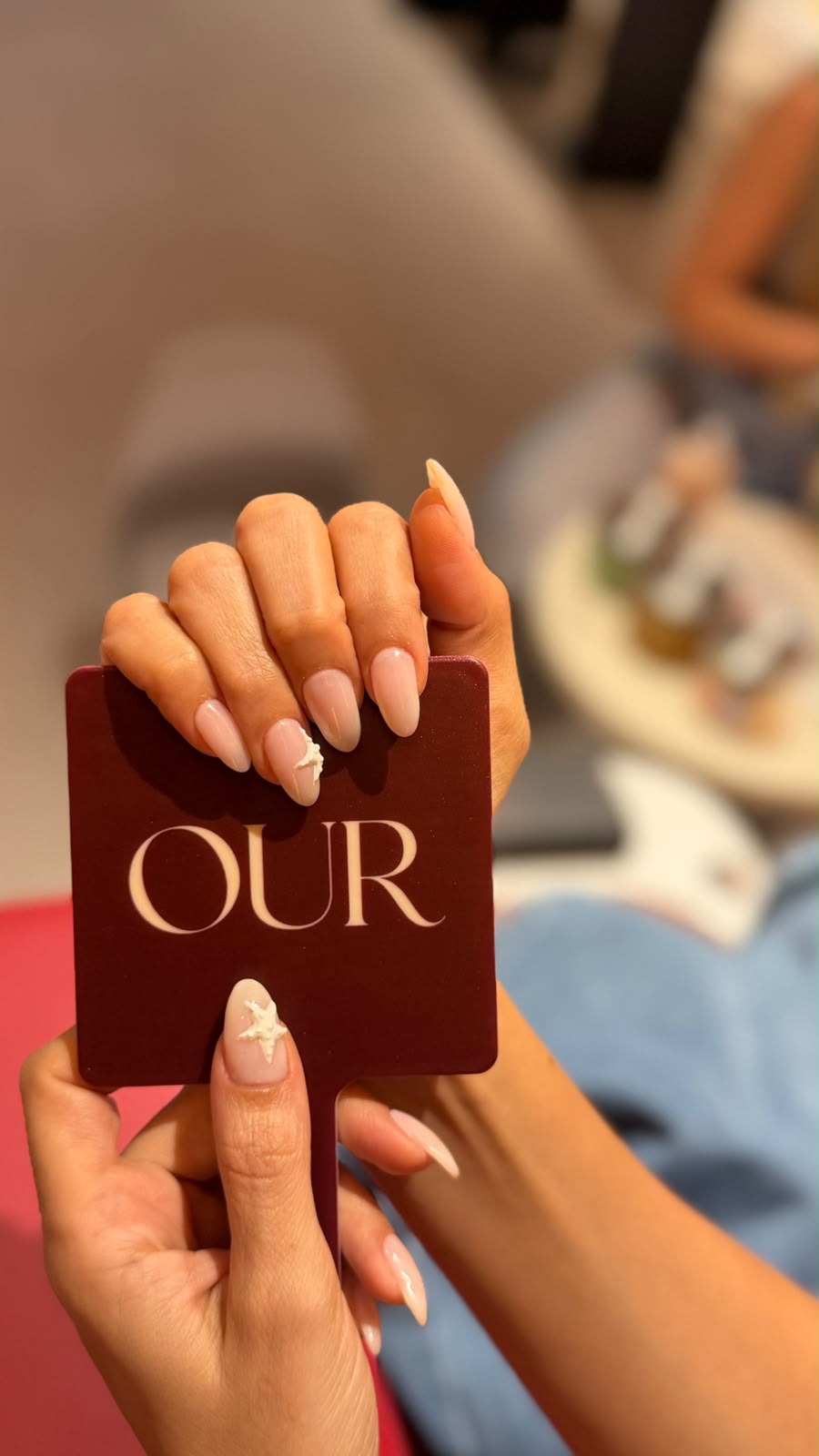 Client holding a burgundy OUR hand mirror showing nude almond nails with 3D floral art