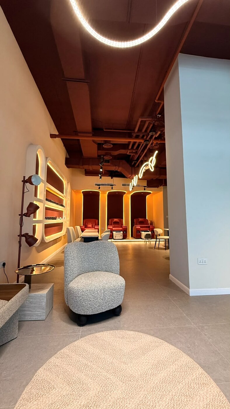 Warmly lit salon lounge with neon detail, boucle armchair and pedicure alcoves beyond